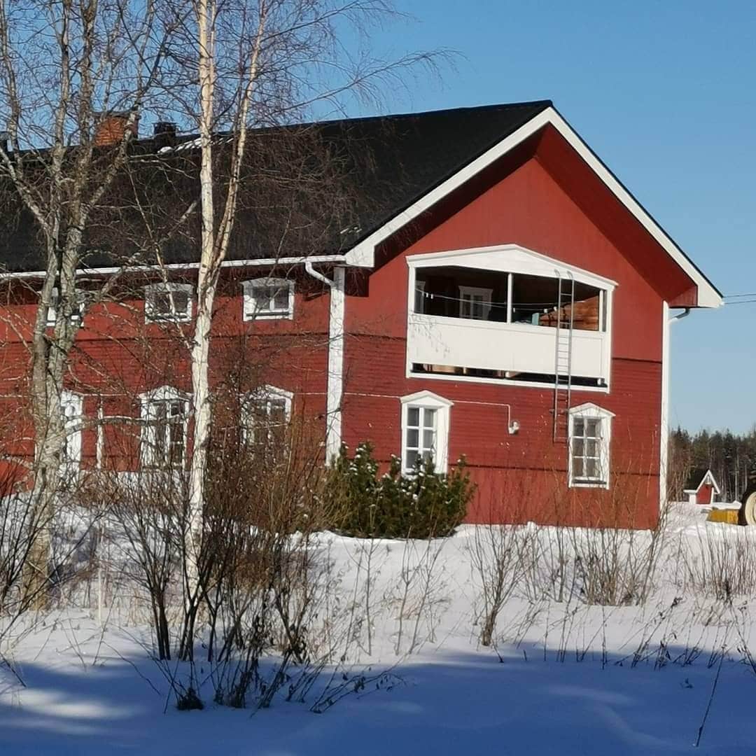 A charming red-painted building features a balcony extending from the upper floor. Snow blankets the ground, and trees with bare branches surround the structure, enhancing the serene winter landscape. Multiple windows allow natural light into the interior spaces.