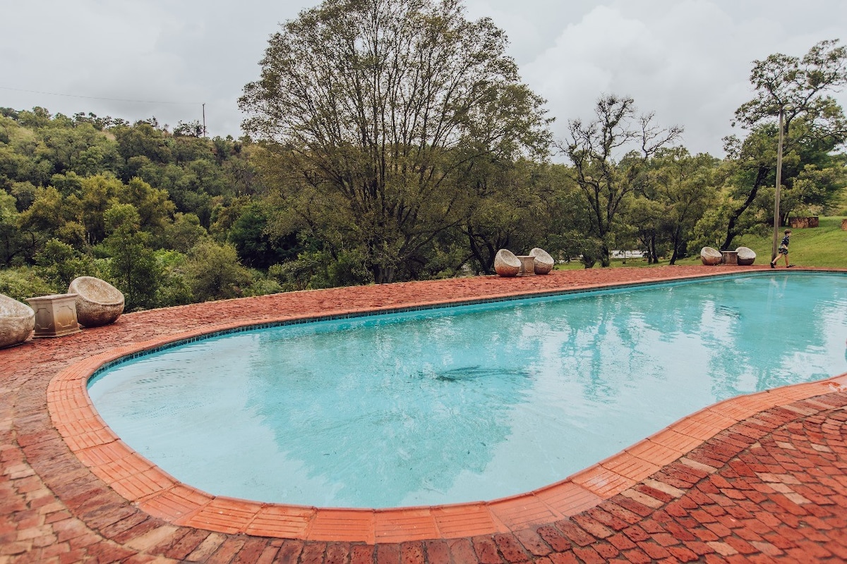 A rectangular pool with clear blue water is surrounded by red brick paving. Natural vegetation and trees create a serene backdrop, with additional seating arranged at the pool's edge, inviting relaxation in a peaceful outdoor environment.