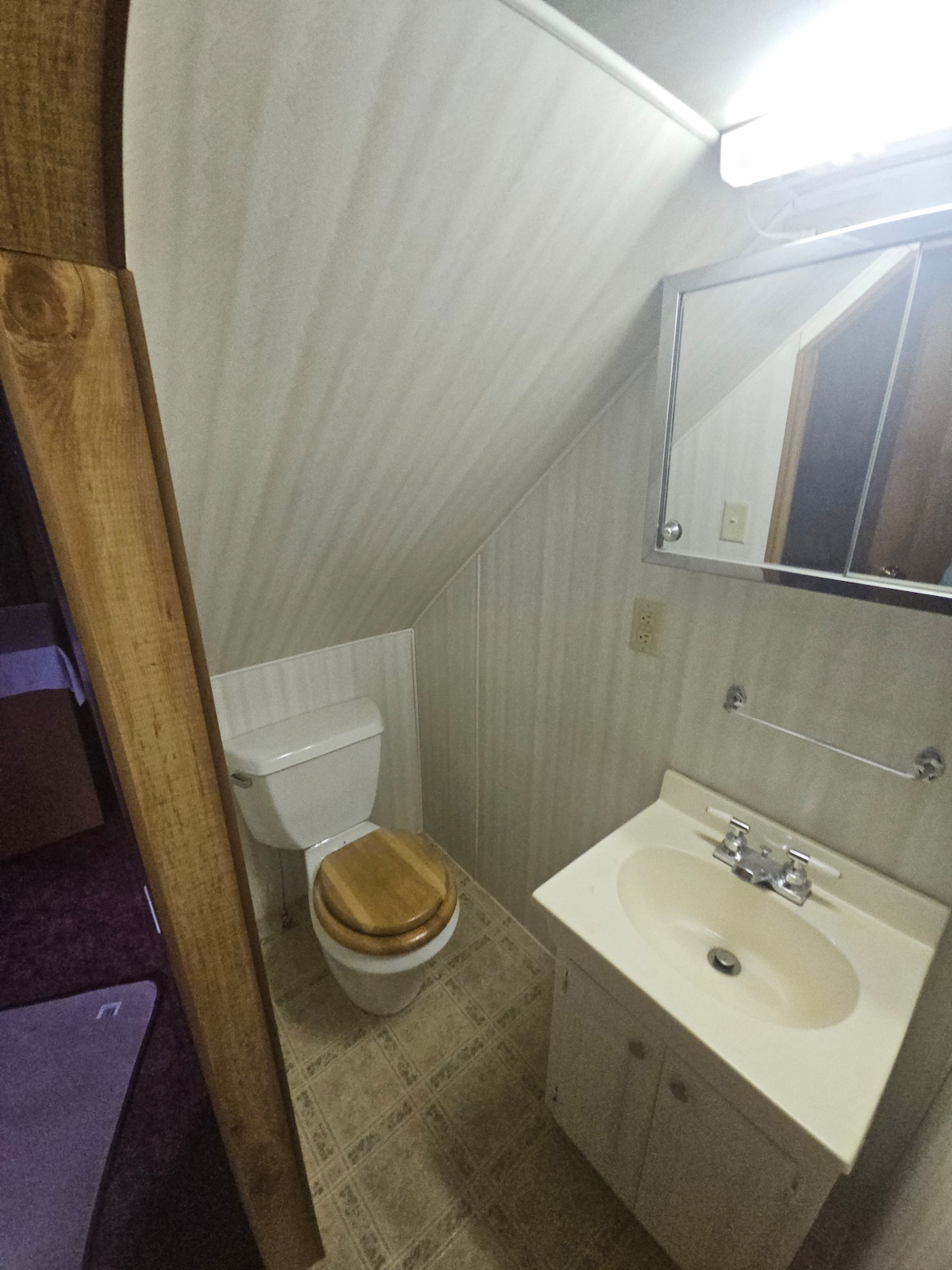 A compact bathroom features an angled ceiling, with a wooden toilet seat and a small sink beneath a mirrored cabinet. Neutral walls and tiled flooring create an understated look, and a towel rack is mounted above the sink.