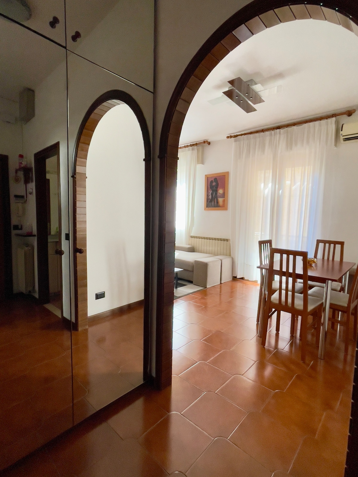 An open living space exhibits an inviting layout featuring an archway entrance. A dining area with wooden chairs and a table is visible, alongside a cozy seating arrangement. Natural light filters through sheer curtains, illuminating the terracotta tile flooring.