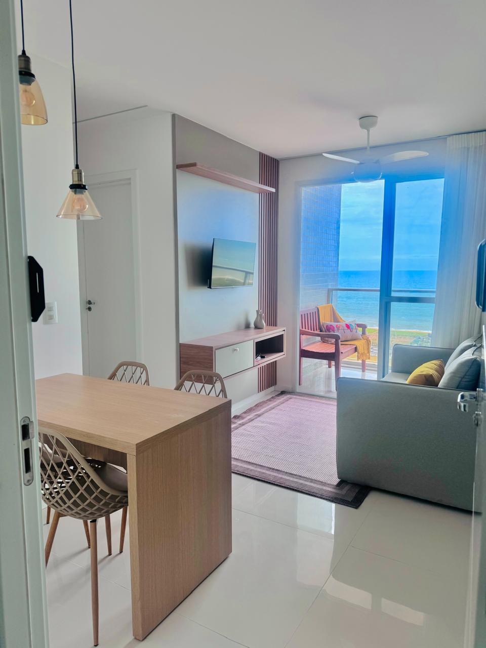 A bright living area features a small dining table and stylish chairs, with a soft gray sofa providing comfort. Floor-to-ceiling windows allow natural light and scenic ocean views. A modern flat-screen television is mounted on the wall, enhancing the contemporary design of the space.