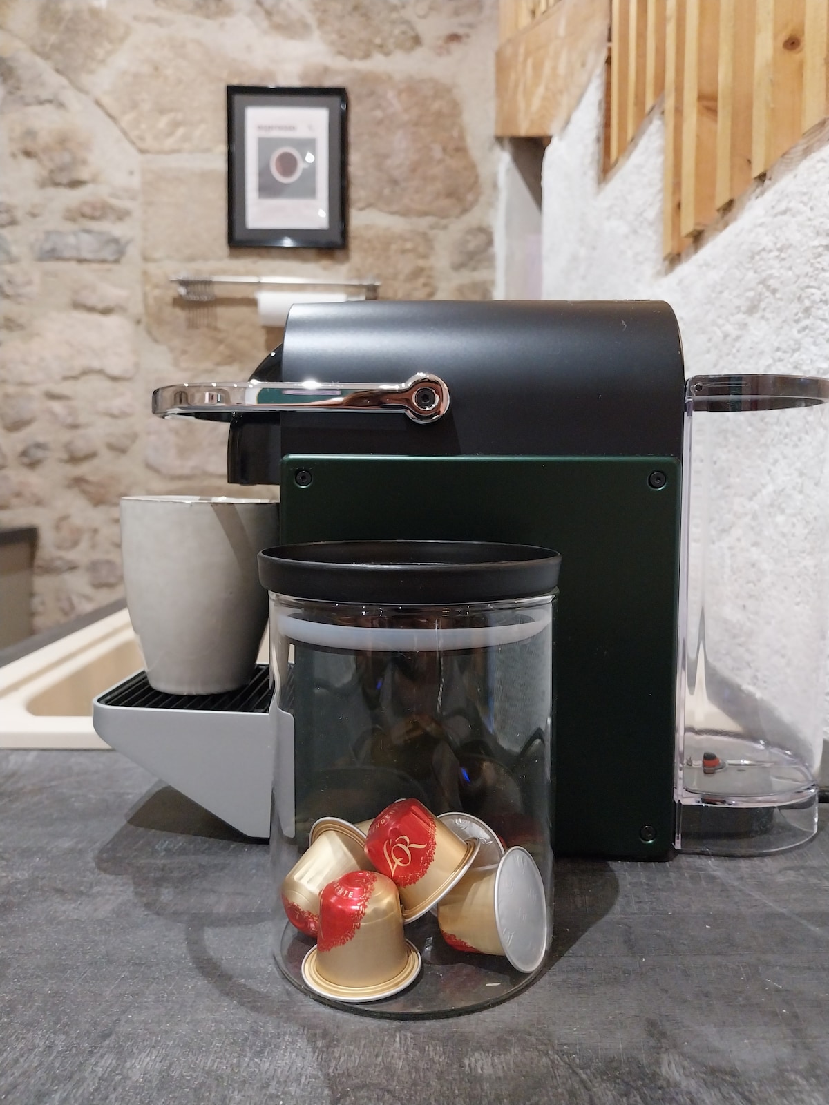 An espresso machine is positioned against a stone wall, accompanied by a sleek glass container filled with coffee capsules. A minimalistic cup stands nearby, with a softly lit background highlighting the cozy kitchen setting.