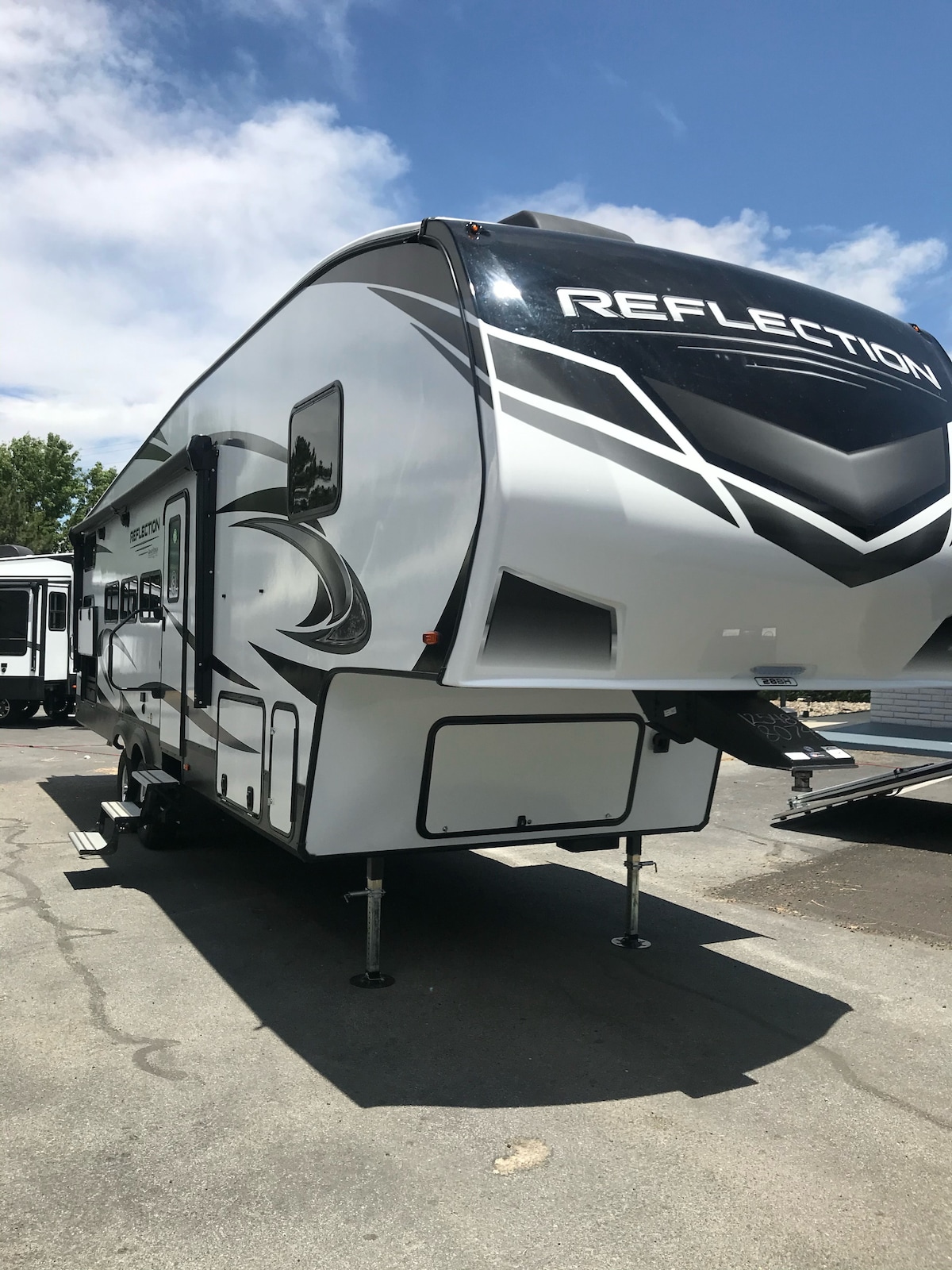 A modern RV is showcased with its sleek exterior design and shiny silver finish. The logo ‘REFLECTION’ is prominently displayed on the side. The vehicle is positioned on level ground, with stabilizing jacks visible, under a clear blue sky.