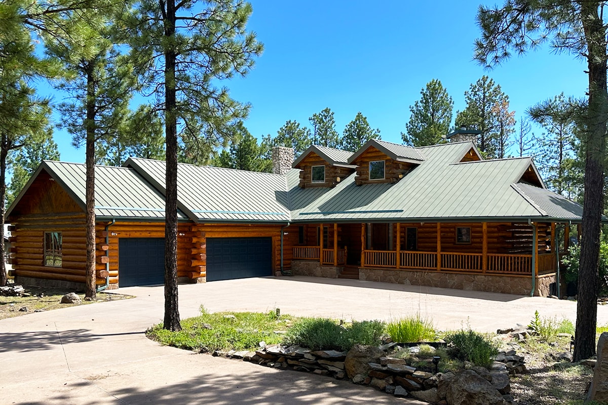 The log cabin exterior is framed by tall pine trees, featuring a distinct sloped metal roof. A spacious driveway is visible, leading to a double garage. The wrap-around porch invites relaxation, showcasing the cabin's rustic charm and integration with the surrounding landscape.