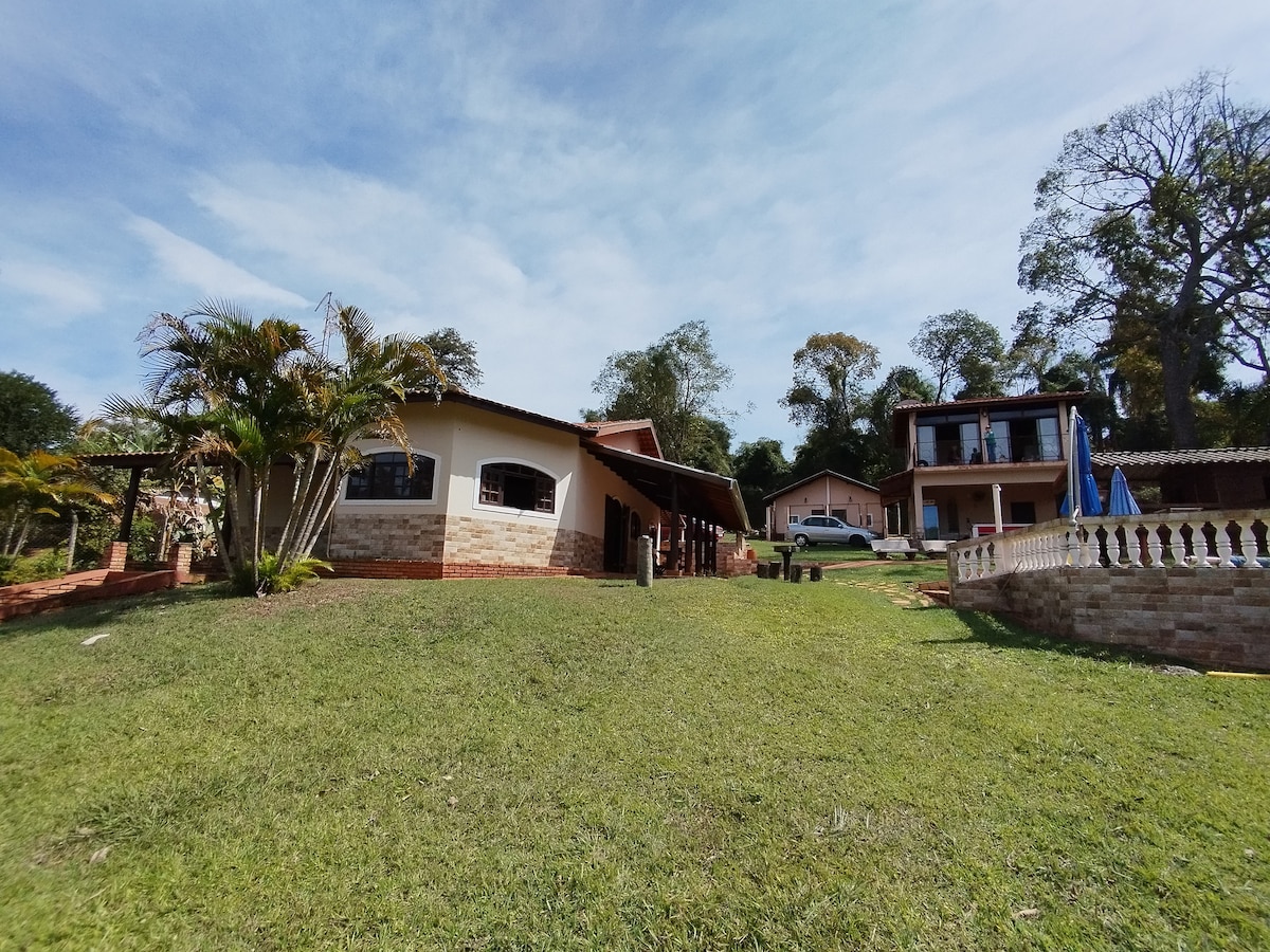A spacious outdoor area is presented, with a well-maintained lawn surrounded by various trees. Two distinct houses are visible, showcasing different architectural styles, with one featuring a porch and large windows, inviting natural light.