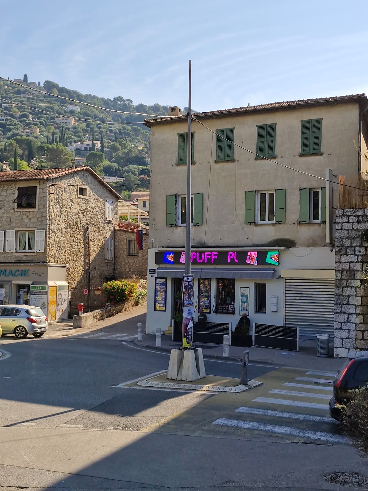 A view of a street corner featuring a light-colored building with green shutters and a signage displaying the word 'PUFF' in bright colors. Stone and stucco buildings are visible in the background, with a hillside covered in greenery rising in the distance. A traffic pole is situated in the foreground.