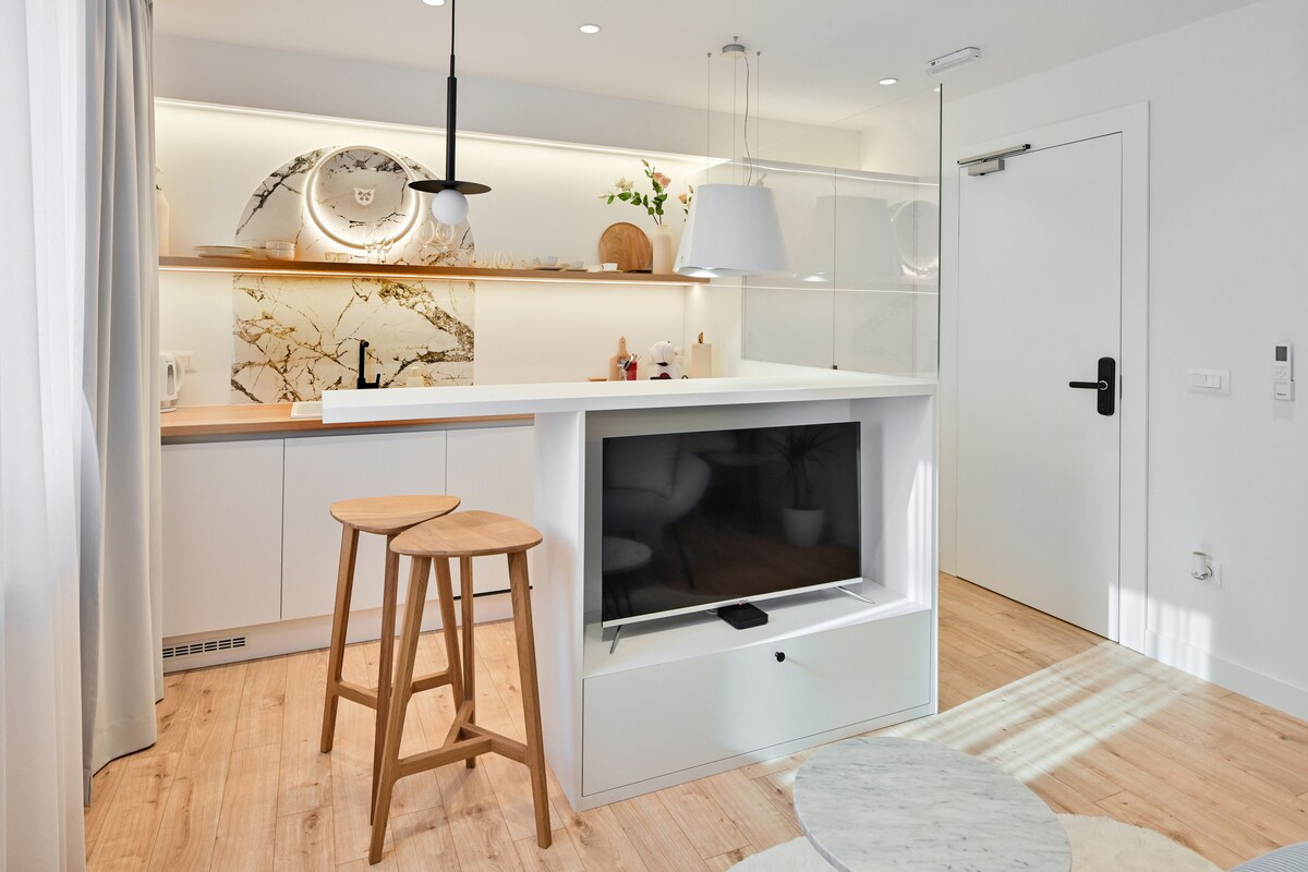A modern kitchenette is showcased with sleek cabinetry, an elegant countertop, and a decorative backsplash. Two wooden stools are positioned at a breakfast bar. Natural light enters through a nearby window, highlighting the minimalist design and soft color palette.