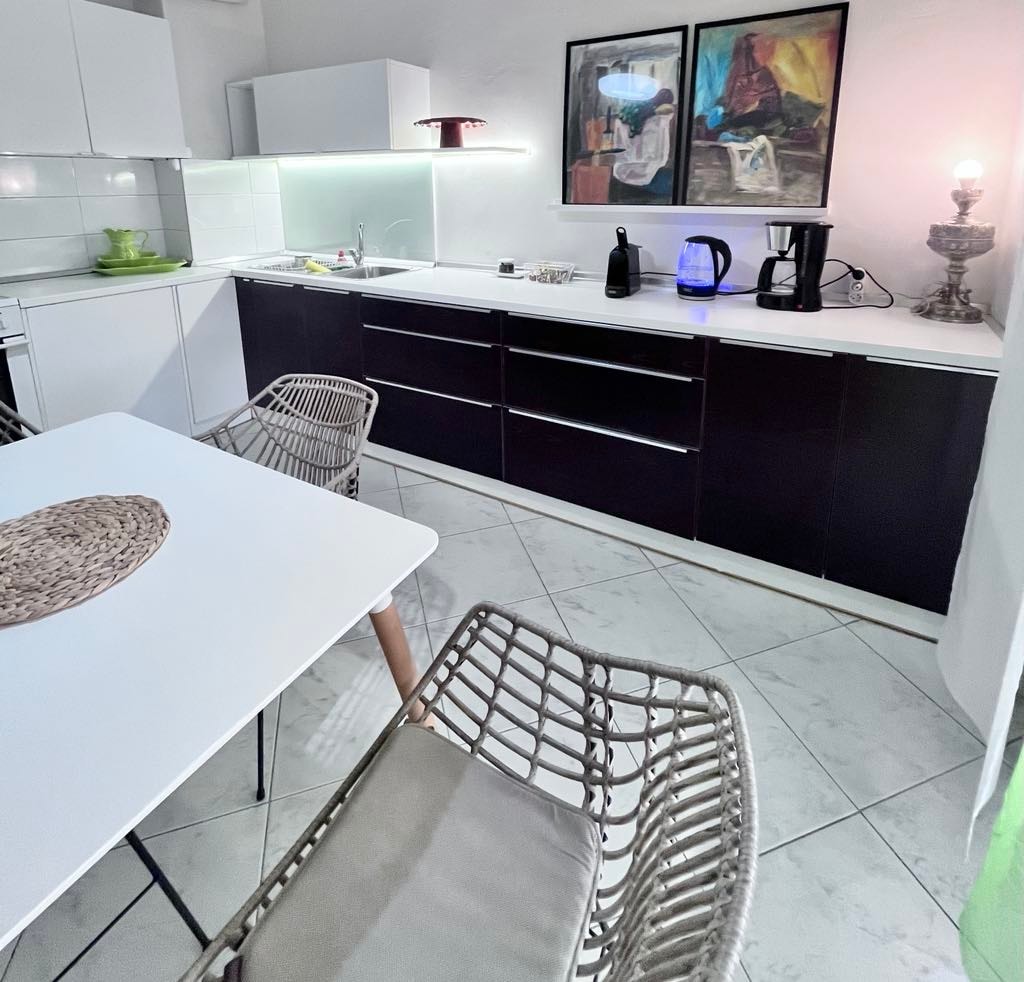 A modern kitchen features sleek cabinetry in dark tones, complemented by a bright countertop. A round dining table is set with a woven placemat, surrounded by two stylish chairs. Art pieces adorn the wall, while appliances include a kettle and coffee maker on the counter.