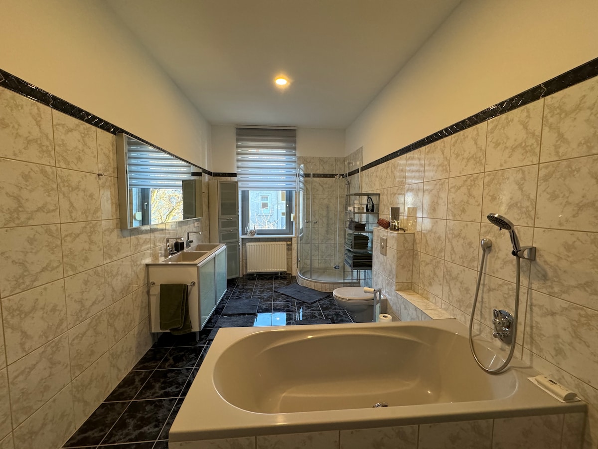 A spacious bathroom features light-colored tiles and an elegant bathtub. Directly opposite, a glass shower enclosure is visible. Two large windows allow natural light to enter, and modern fixtures are included, along with a stylish sink and ample counter space.