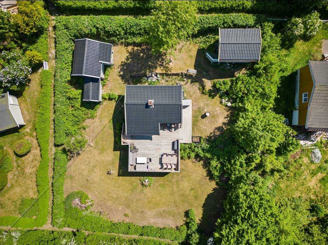 An aerial view captures a charming cabin surrounded by lush greenery. The structure features a dark roof and a spacious outdoor deck with seating. Two smaller annexes are visible in the garden, which is neatly bordered by hedges and pathways.