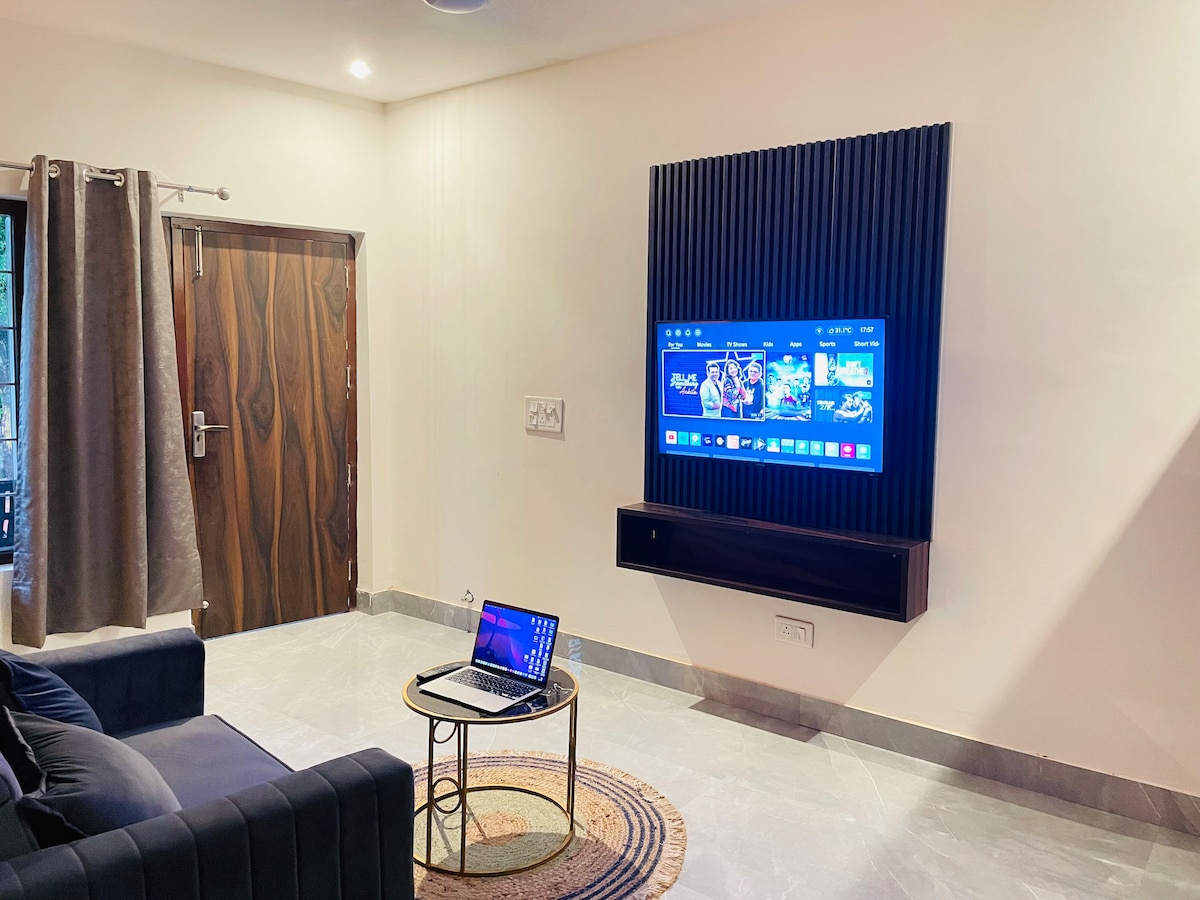 A modern living area features a dark sofa, a round coffee table, and a wall-mounted television displaying a vibrant interface. Natural light enters through a nearby window covered by curtain panels. A stylish door with a wood finish adds character to the space.