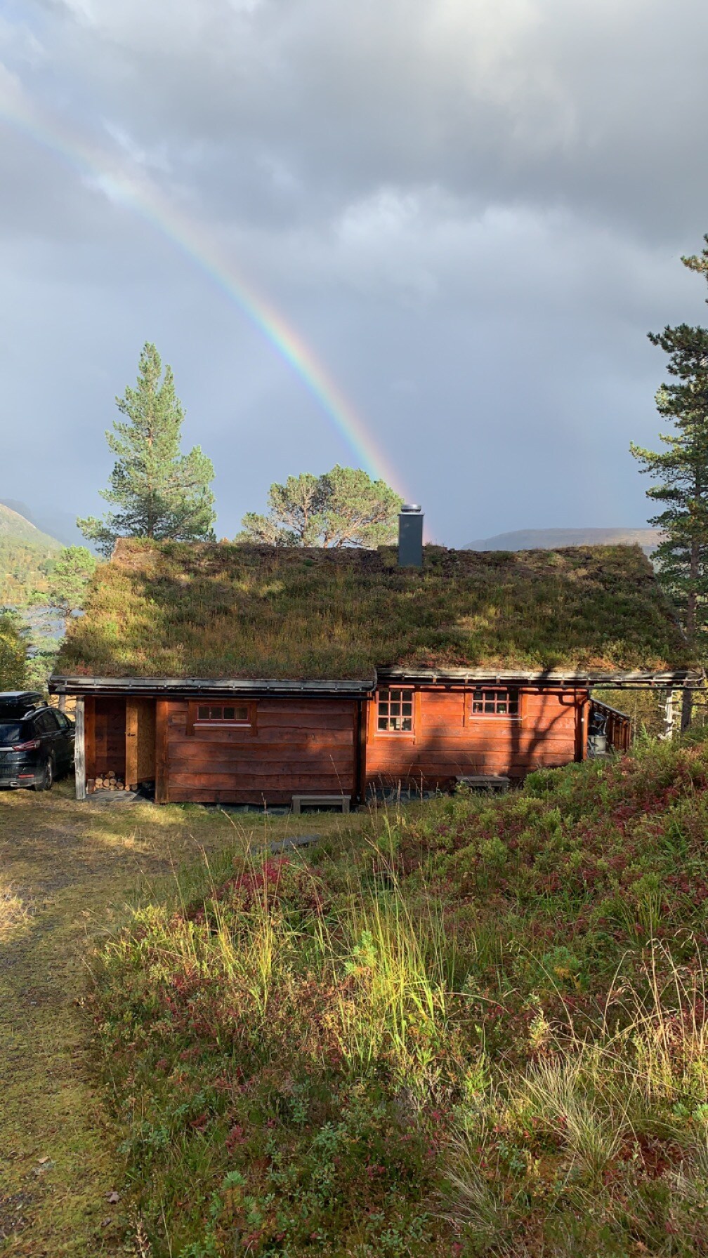 A wooden cabin with a grass-covered roof is positioned among greenery. A soft rainbow is visible in the sky above, adding a touch of color to the serene landscape. The cabin features multiple windows that reflect its rustic charm.