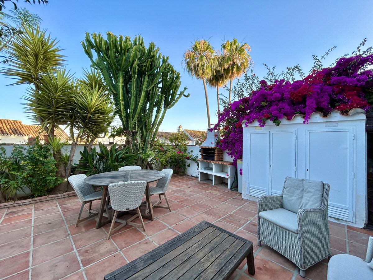 A private terrace is shown, featuring a round dining table surrounded by four wicker chairs. Bougainvillea and various tropical plants provide vibrant greenery, while a BBQ area is visible under a wooden awning. The terracotta floor adds warmth to the outdoor space.