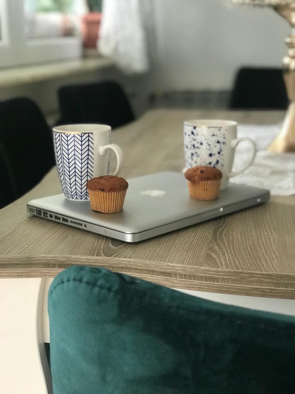 An inviting dining table features a wooden surface with two mugs and two muffins placed on a silver laptop. Soft, green upholstery and dark chairs are visible, with natural light illuminating the space through nearby windows.