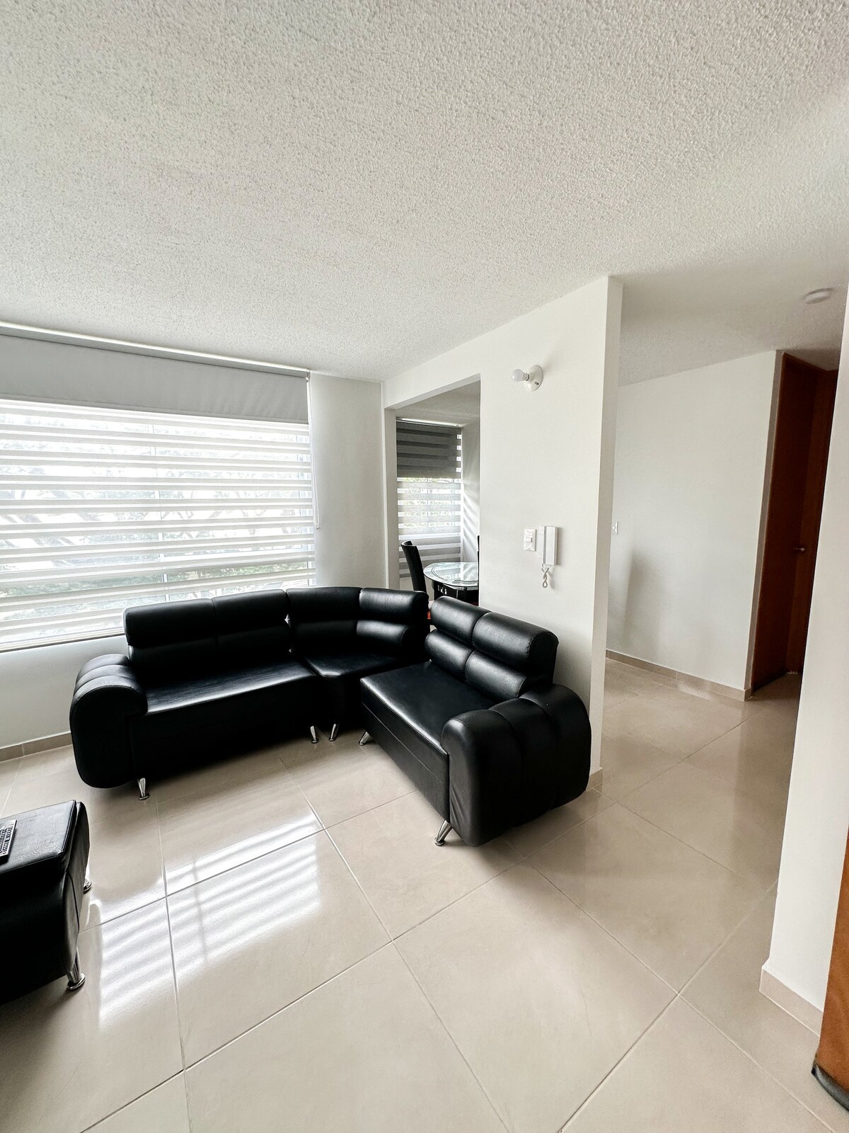 A modern living area features a black sectional sofa positioned against a wall. Natural light enters through large windows adorned with horizontal blinds, reflecting softly on the polished floor. A glimpse of a mirrored wall adds a sense of spaciousness to the room.
