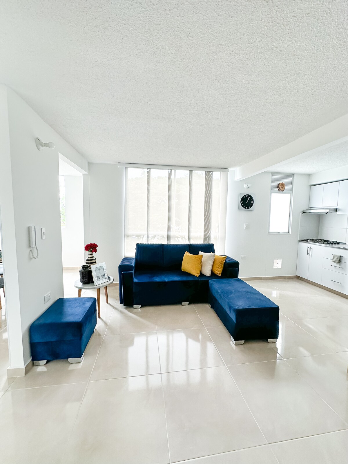 A modern living area features a blue sectional sofa with yellow cushions, paired with matching ottomans. Large windows allow natural light to fill the space, while a wall clock and minimalist kitchen are visible in the background, creating a bright and inviting atmosphere.