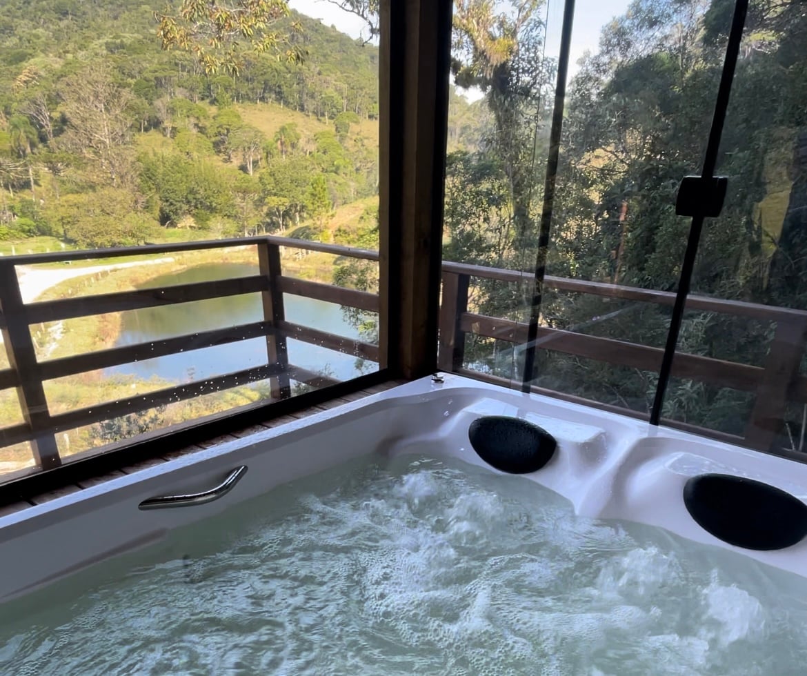 A luxurious hot tub is situated by large glass windows, offering views of lush greenery and a serene body of water outside. The bubbling water invites relaxation, while natural light fills the space, creating a tranquil atmosphere.