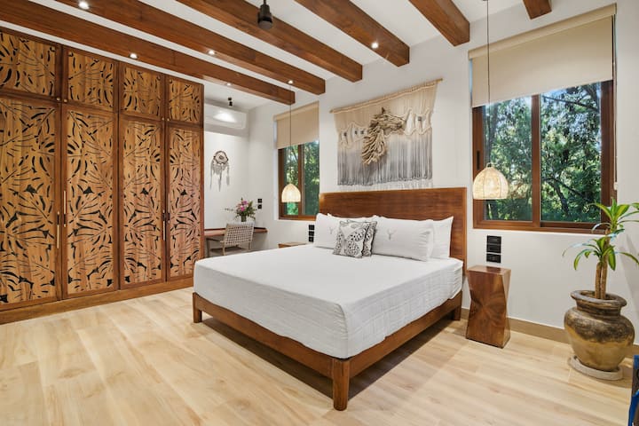 Con una mezcla de texturas que combinan escultura de madera y macramé personalizado, el sereno dormitorio tamaño king de arriba cuenta con vistas a la selva, iluminación al atardecer, amplio espacio en el armario y un escritorio