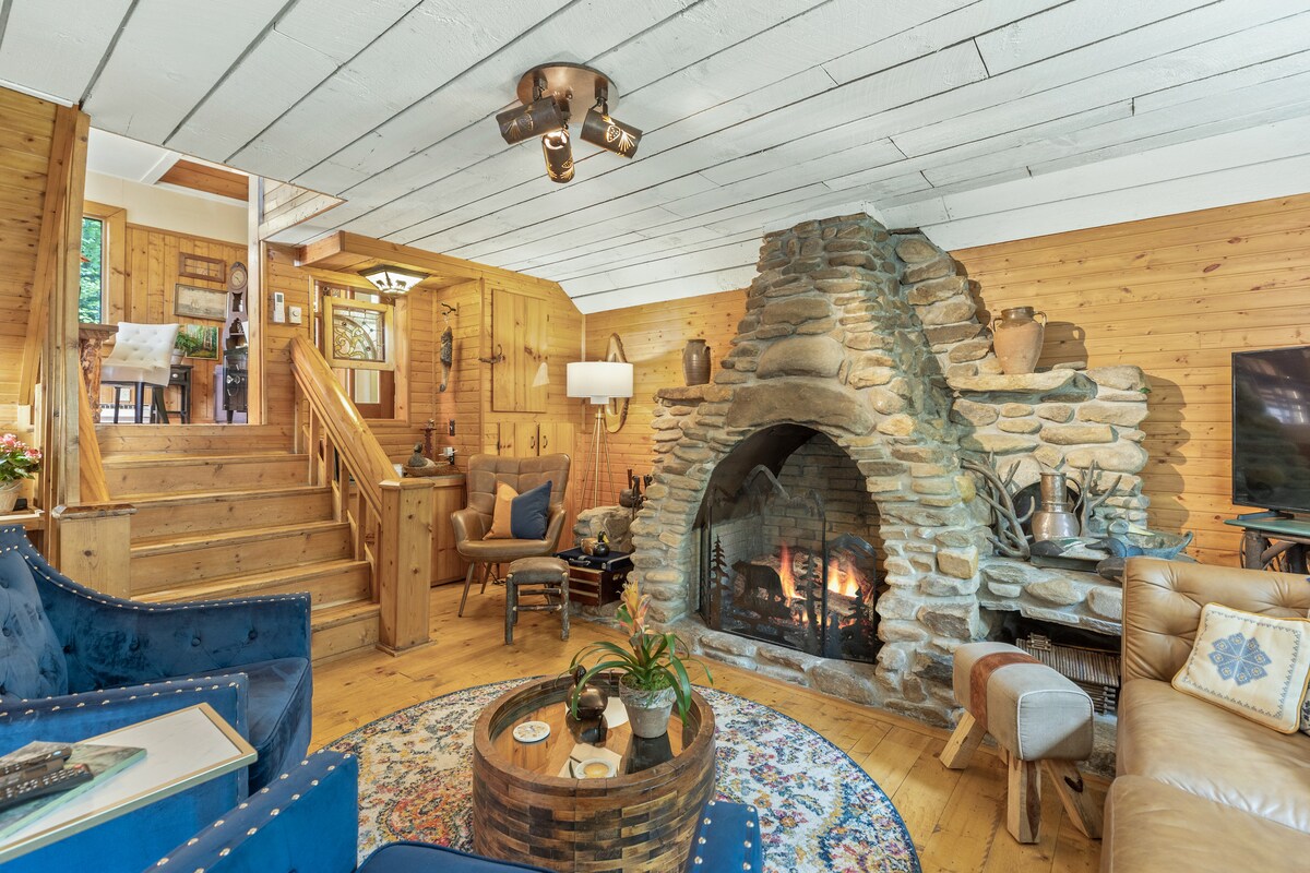 The great room features a custom stone fireplace as the central focus, surrounded by comfortable seating options. Wood-paneled walls create a cozy ambiance, while a circular rug adds warmth to the space. A staircase provides access to upper levels, enhancing the sense of structure.