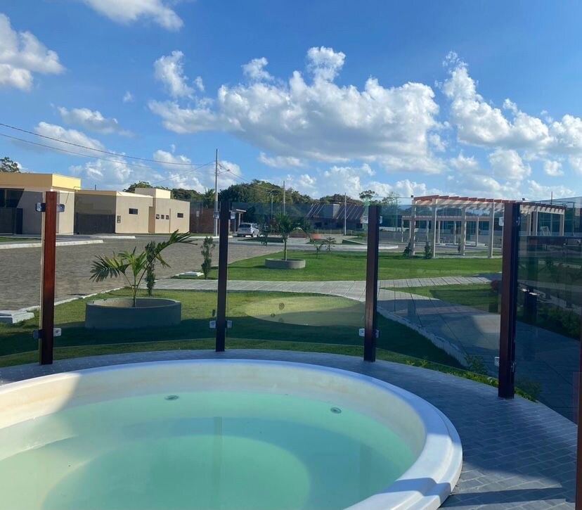 A circular hot tub is positioned in the foreground, with clear water visible. Beyond the tub, an expansive green area is framed by an elegant glass barrier, with palm trees dotting the landscape and buildings in the background under a bright, blue sky.