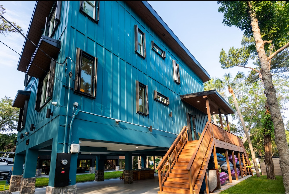 A modern lagoon house features a striking blue exterior and a spacious wooden staircase leading to the entrance. Large windows allow natural light to filter through, surrounded by lush greenery and palm trees. The ground level provides ample parking and outdoor seating options.
