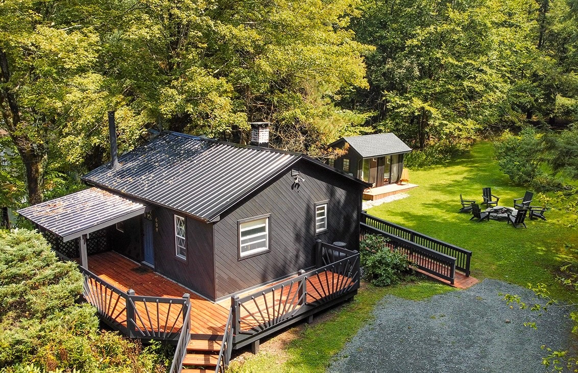 The exterior of a cozy black cottage is surrounded by lush greenery, featuring a wooden deck with railings and stairs leading to the entrance. A separate outdoor room is visible in the yard, along with a fire pit and several chairs arranged for relaxation.