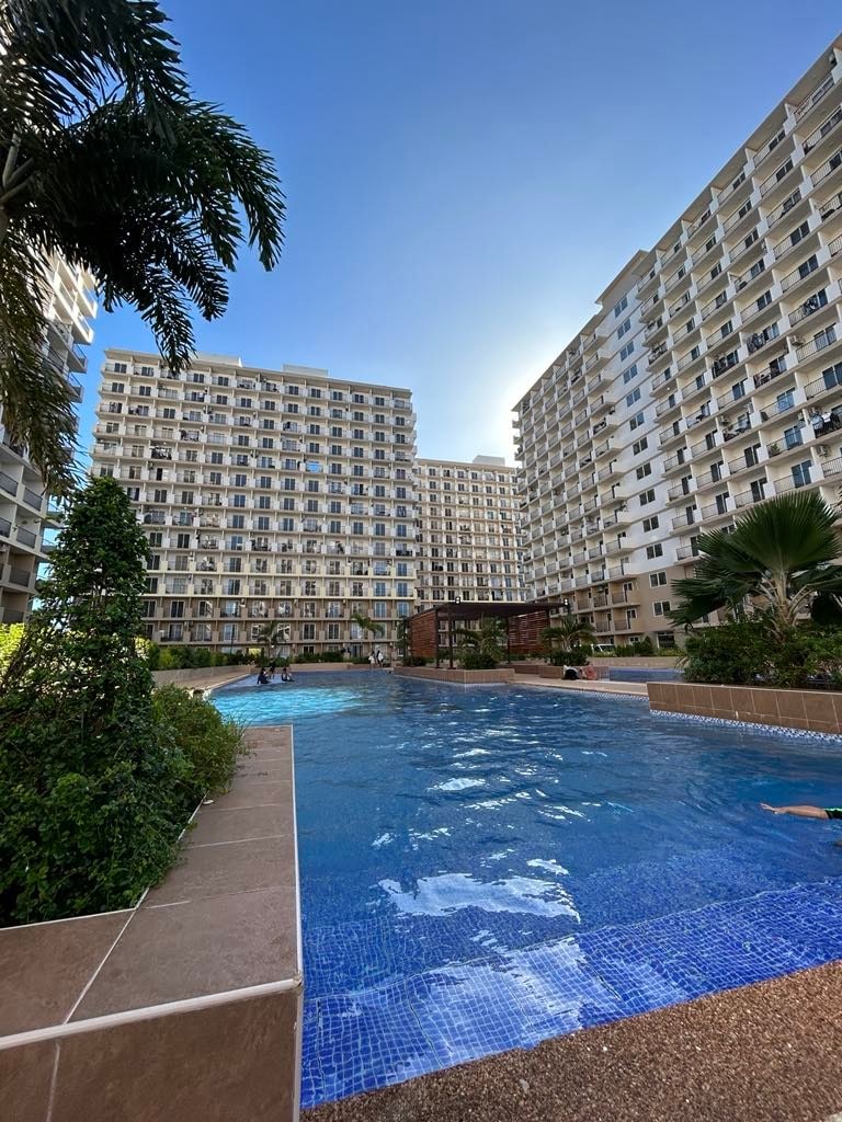 A spacious swimming pool is situated within a landscaped courtyard, surrounded by several multi-story buildings. Lush greenery borders the pool area, with the sunlight reflecting off the water's surface, creating a serene ambiance.