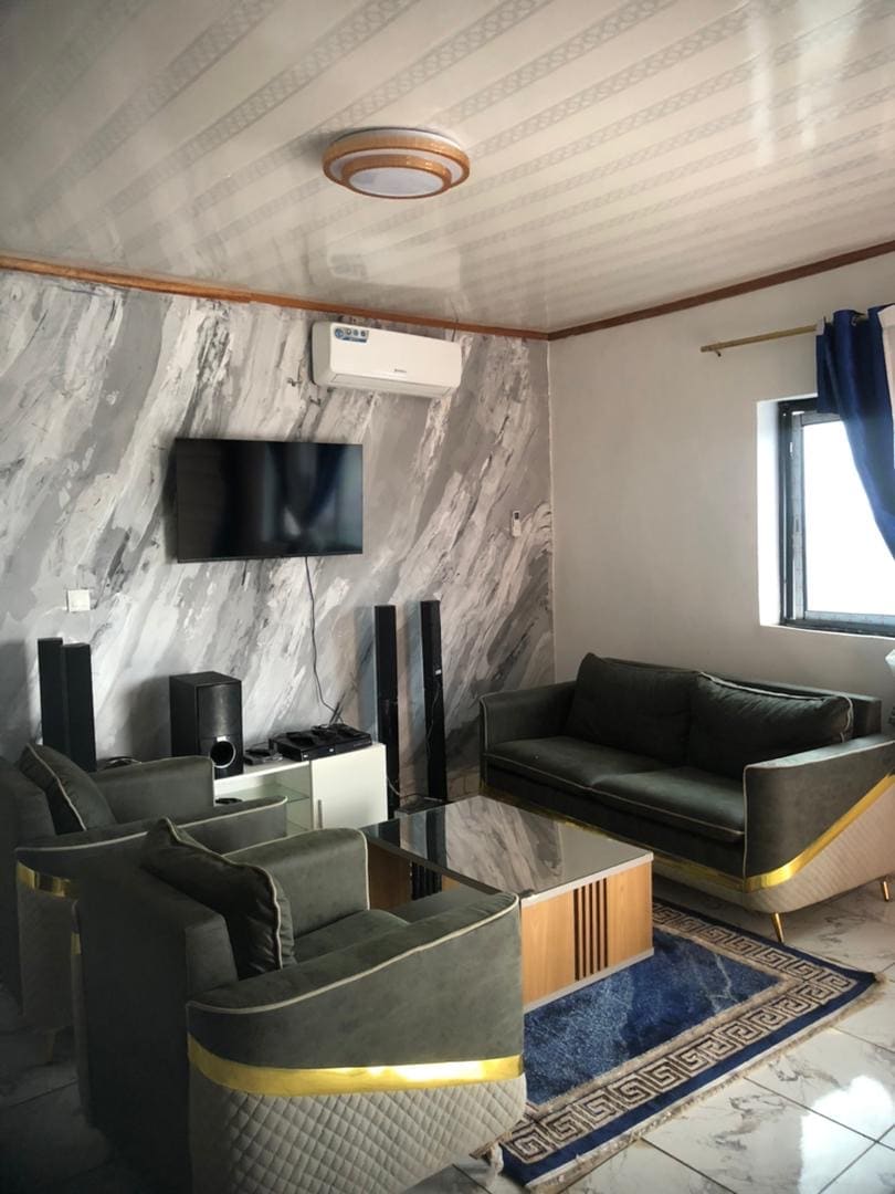A modern living area features a sleek television mounted on a marbled wall. Two comfortable sofas are positioned around a glass-topped coffee table, and stereo speakers flank the space. Natural light enters through a window, elegantly dressed with blue curtains.