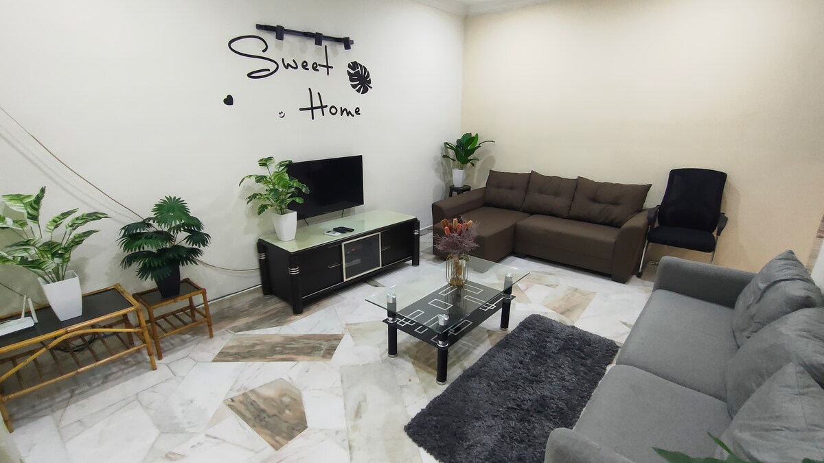 A welcoming living area features comfortable seating arrangements with a brown sectional sofa and a single armchair. A coffee table with a decorative vase sits centrally, while wall-mounted plants add a touch of greenery. The phrase 'Sweet Home' is displayed on the wall, emphasizing a cozy atmosphere.