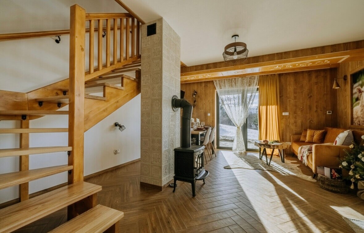 A spacious living area features a wooden staircase leading to an upper level. Natural light filters through large windows, illuminating the warm wooden walls. A cozy seating arrangement is present, complemented by a decorative stove and subtle textile accents.