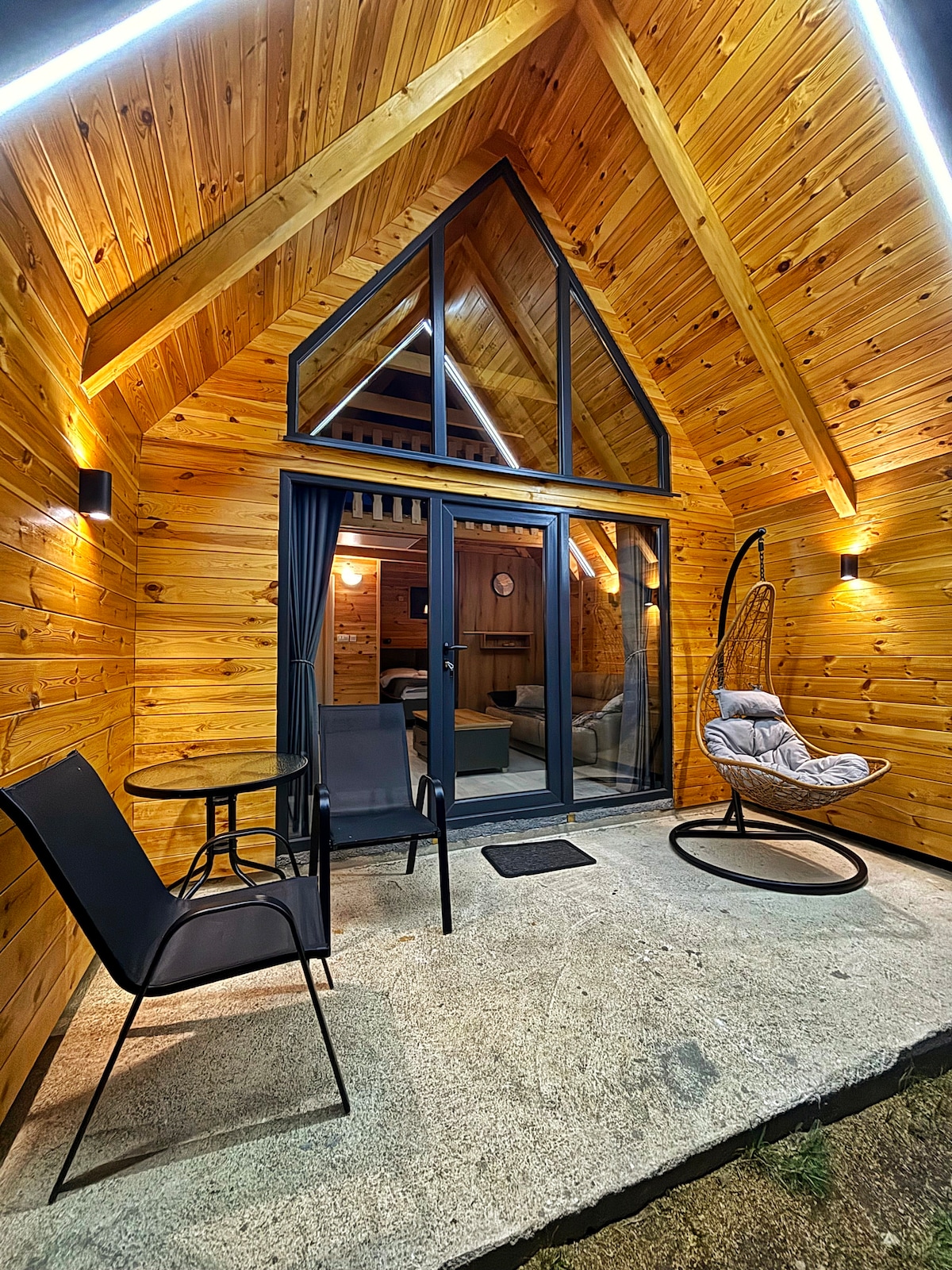 A modern cabin exterior showcases natural wood paneling and an A-frame roof design. A glass door leads to the interior, flanked by a hanging chair and a small table with two chairs. Subtle lighting enhances the welcoming ambiance.