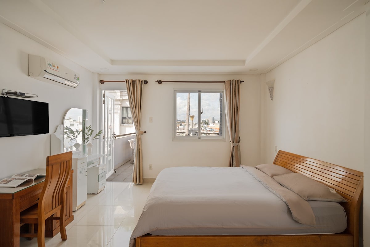 A well-lit bedroom features a spacious bed adorned with a light gray comforter. A wooden table and chair are positioned nearby, accompanied by a mirror. Natural light filters through the window, which is framed by neutral-toned curtains, enhancing the serene ambiance.