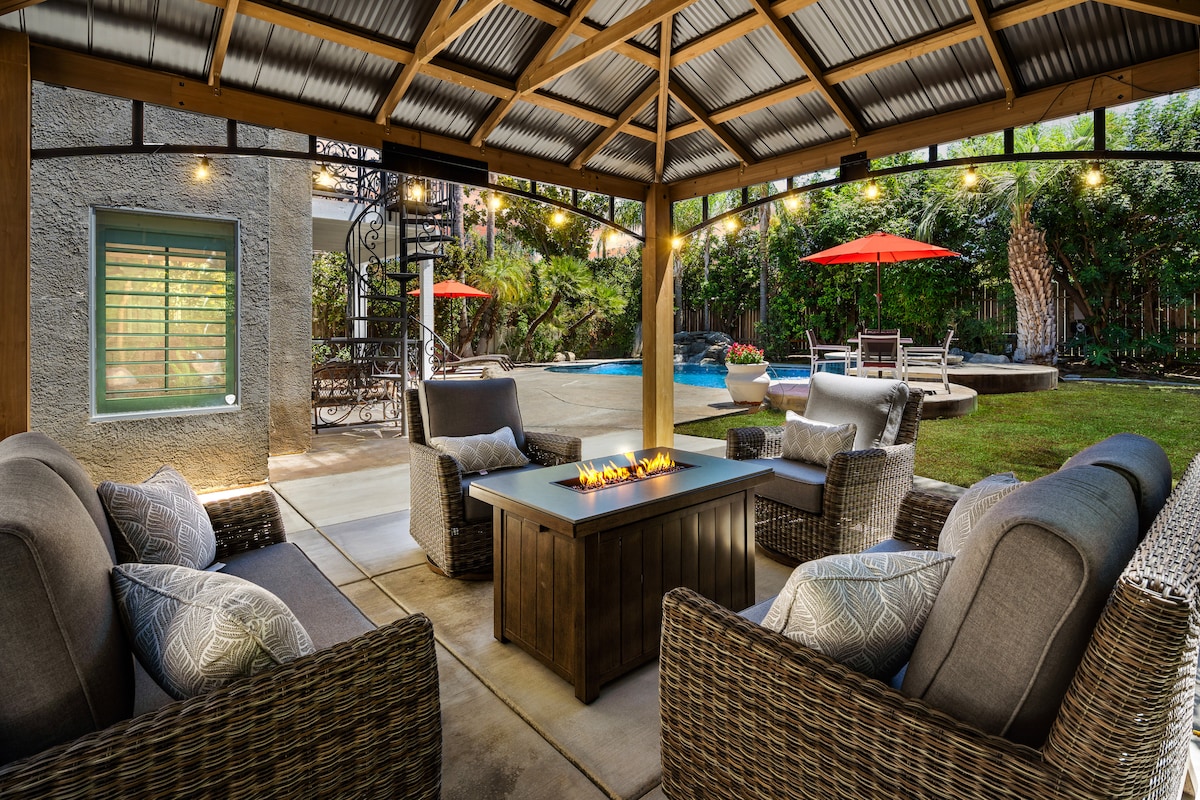 A covered gazebo features comfortable seating arranged around a sleek fire pit. Soft lighting from string lights enhances the inviting ambiance, while the pool, hot tub, and outdoor lounge chairs are visible in the landscaped backyard beyond.