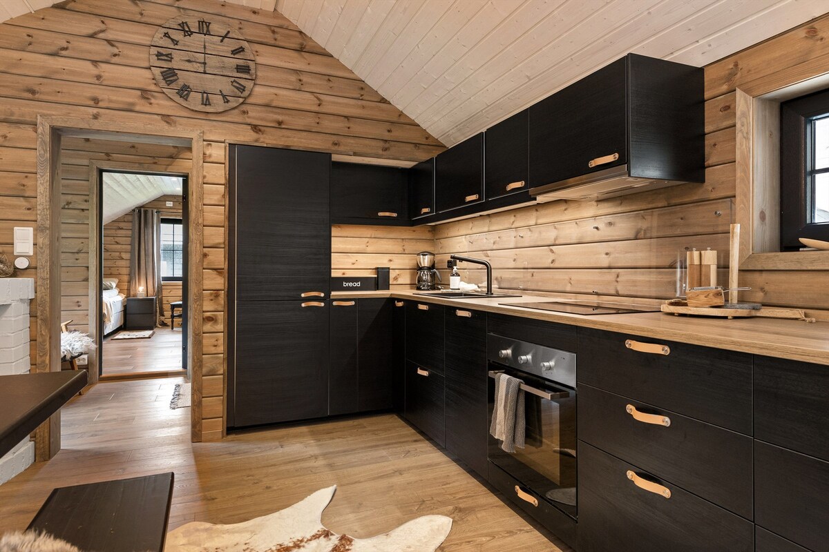 A modern kitchen is featured within a cozy wooden cabin, showcasing sleek black cabinetry with wooden handles. A stainless-steel oven is positioned alongside a minimalist sink. Natural light floods the space, highlighting the warm wood textures of the walls and ceiling.