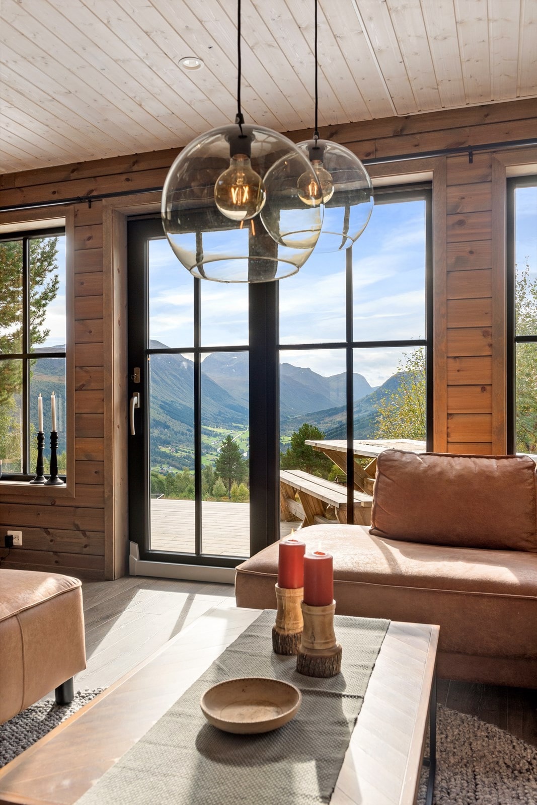 The interior space features large glass doors that open to stunning mountain views, allowing natural light to fill the room. Soft leather seating surrounds a wooden coffee table, accented by decorative candles and a bowl. The warm wooden walls enhance the cozy ambiance.
