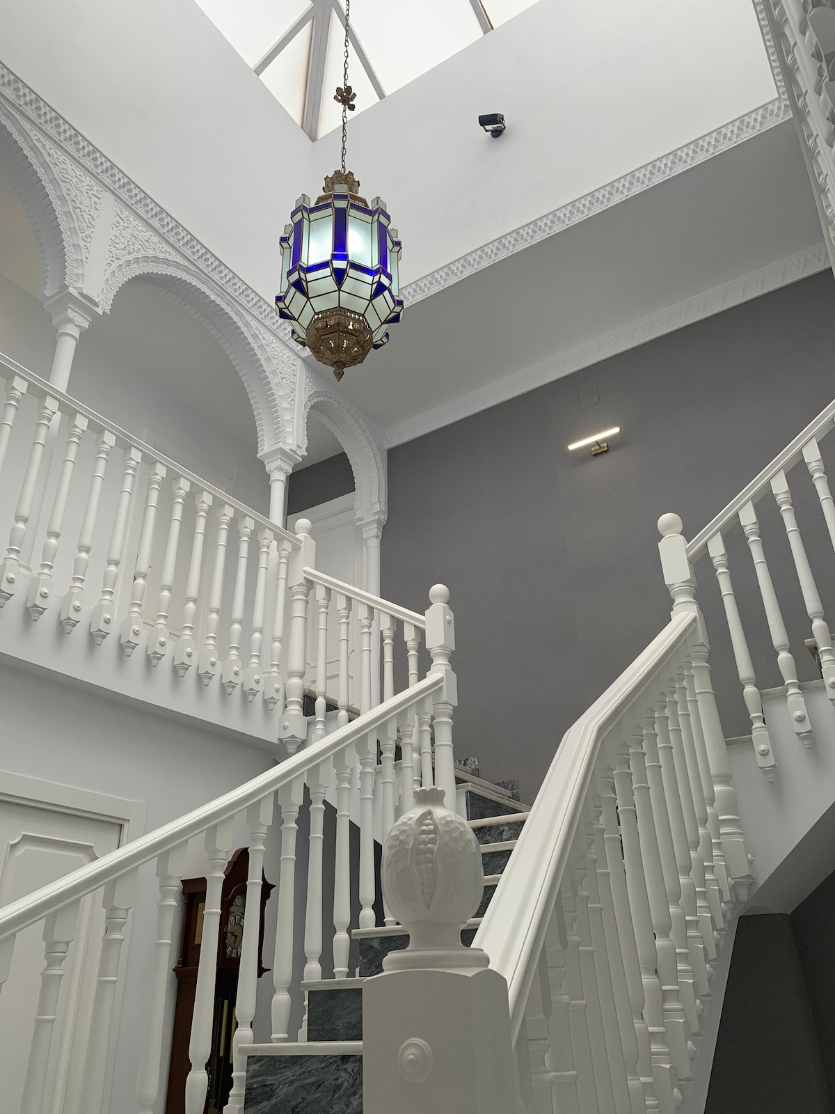 A grand staircase is flanked by intricate white railings, leading to a brightly lit upper level. An ornate lantern hangs above, casting a soft glow. The walls feature detailed arches, contributing to the villa's architectural elegance.