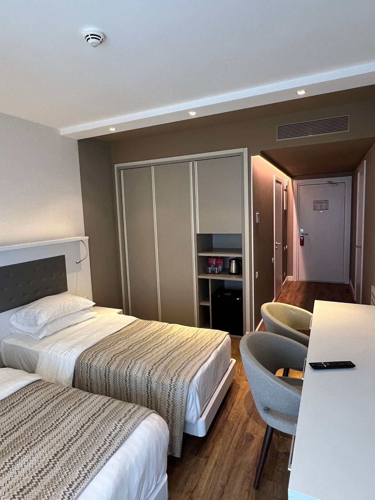 A cozy studio space features two beds with light bedding, placed opposite each other. A minimalist desk and seating area are visible, along with built-in cabinetry. Soft natural light fills the room, highlighting the warm wooden flooring and neutral color palette on the walls.