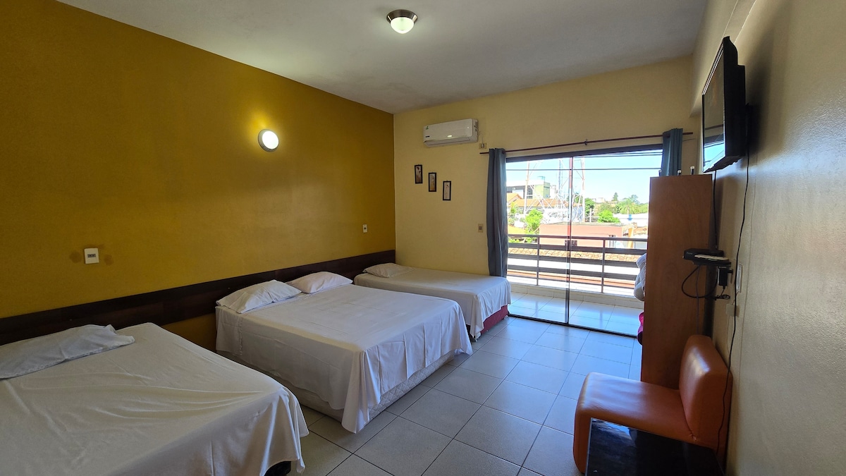 A spacious sleeping area is presented with three beds, each neatly made with white linens. A large window allows natural light to fill the room, highlighting the warm yellow walls. A small seating area comprised of an orange chair and a wall-mounted television is also visible.