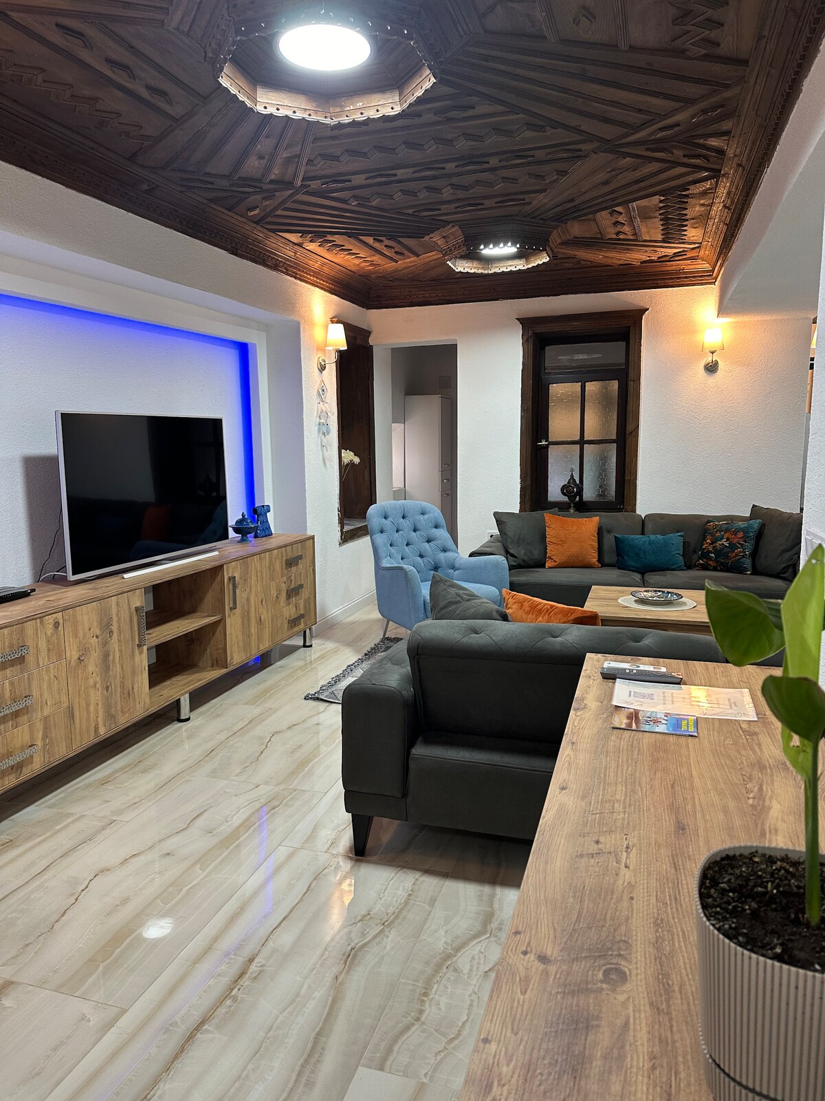 A stylish living area features a combination of plush seating, including a gray sofa and a blue accent chair. A wooden media console holds a flat-screen TV, and decorative cushions add color. Warm lighting highlights the intricate ceiling design, creating a cozy ambiance.