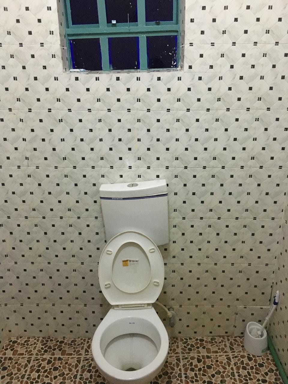 A bathroom is showcased with patterned tiles covering the walls. A flush toilet is centered against the wall, and a small waste bin is positioned nearby. A window permits natural light to enter, framed with a simple green casing.