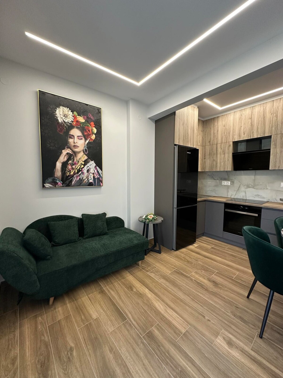 A modern kitchen space features sleek cabinetry with wooden accents. A dark green sofa is complemented by a decorative plant on a small table. An artistic wall painting adds a touch of personality to the room, underlined by soft, ambient lighting.