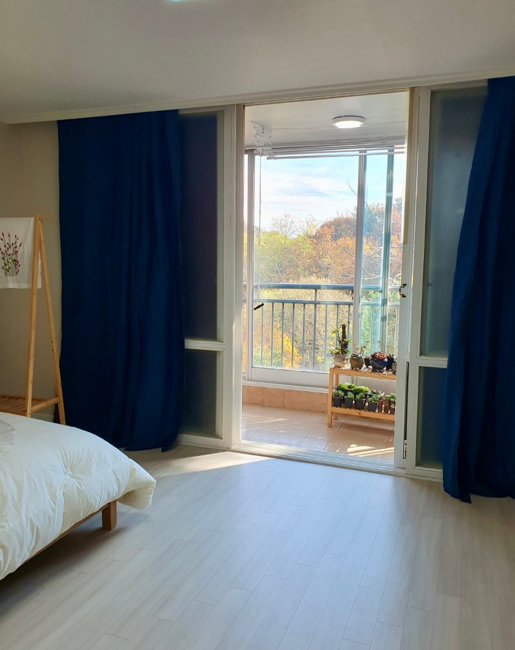 A bright bedroom features a spacious bed draped with a soft blanket. Floor-to-ceiling glass doors open to a balcony, revealing a colorful view of outdoor greenery. Dark curtains frame the windows, while natural light fills the room, enhancing its airy ambiance.