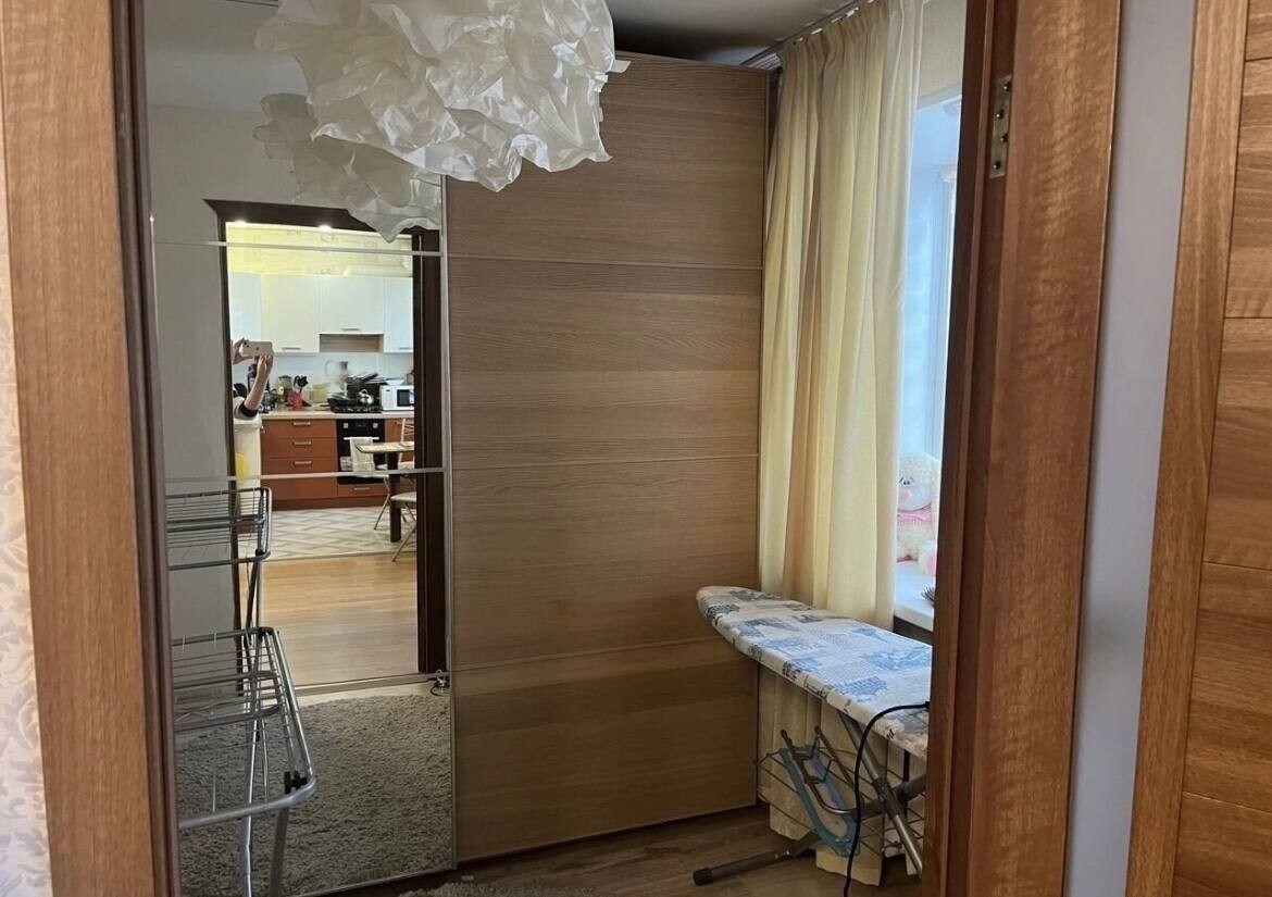 A wooden door provides access to a cozy living area visible in the background. Natural light enters through sheer curtains, enhancing the warm tones of the wooden furnishings. An ironing board is positioned against one wall, with a clothing rack nearby.