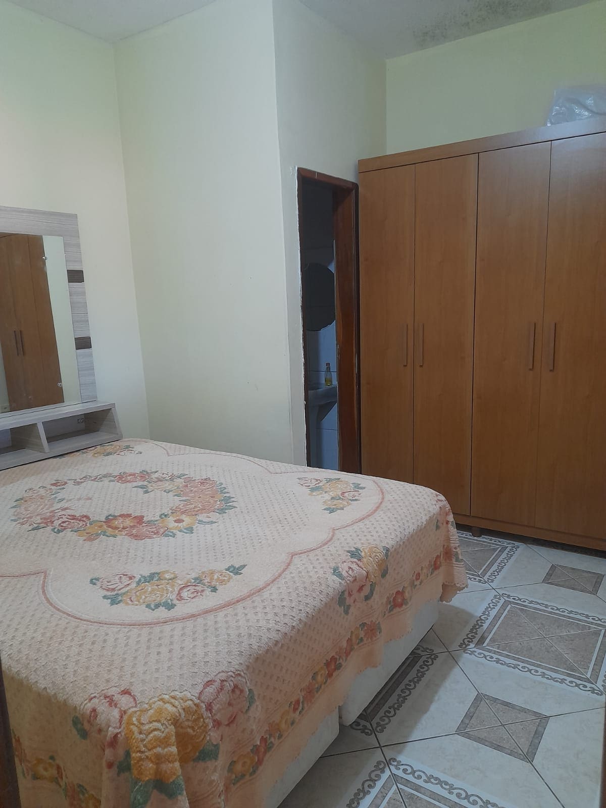 A bright bedroom is furnished with a spacious bed covered by a floral-patterned blanket. A large wooden wardrobe provides ample storage, and a mirror is positioned above a small dresser. The muted yellow walls add a calm ambiance to the room.