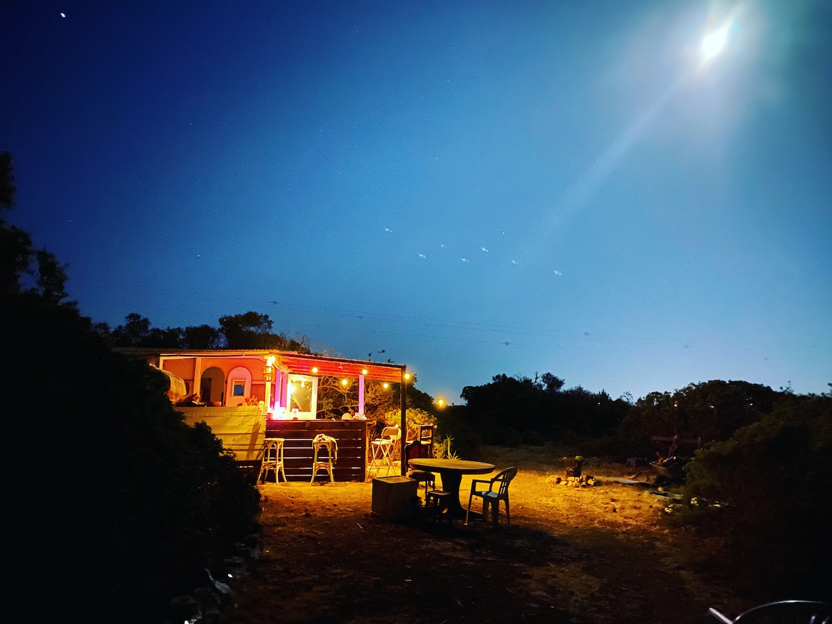 A caravan illuminated by soft lights stands at night, surrounded by nature. Chairs are arranged around a wooden table, creating an inviting outdoor dining area. The bright moon casts a gentle glow over the scene, enhancing the peaceful atmosphere.