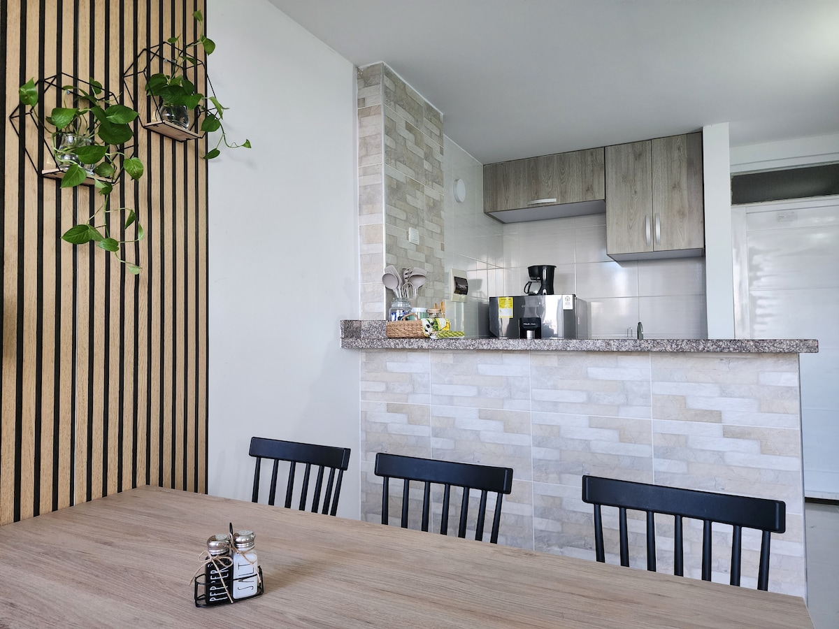 The kitchen area features modern cabinetry with neutral tones and a granite countertop. A coffee maker is positioned beside various kitchen essentials, while a dining table with black chairs provides seating for meals and gatherings. Wall-mounted plants add a touch of greenery.