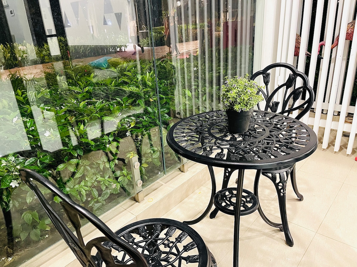 A black metal table with two matching chairs is situated near a glass wall. A small potted plant sits centrally on the table, while a vibrant garden with greenery is visible through the glass, offering a sense of outdoor connection.