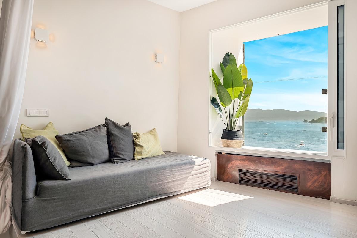 A bright living space features a grey couch adorned with green accent pillows. A large window reveals a stunning view of the sea, framed by a vibrant houseplant. Natural light streams in, enhancing the serene ambiance of the room.