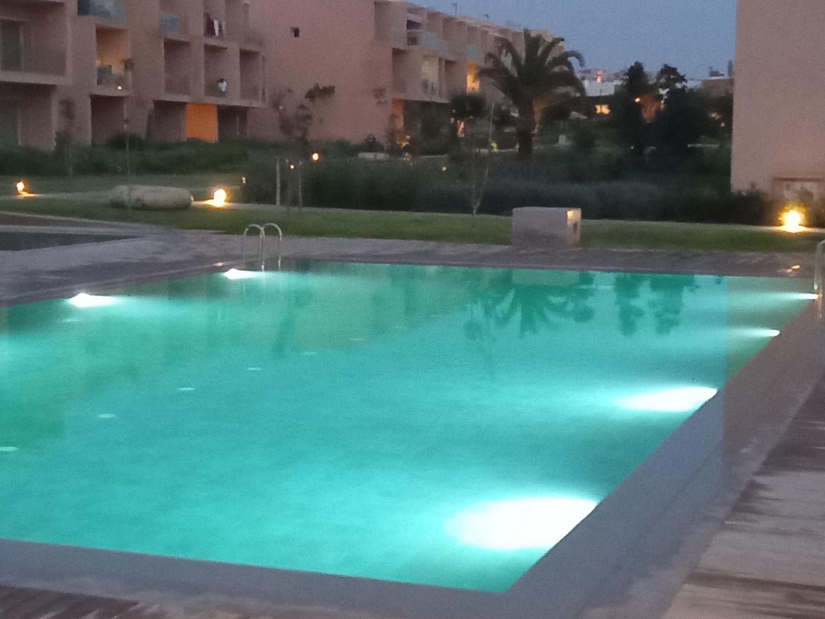 A serene swimming pool is featured, illuminated by soft lights reflecting on the water. Surrounding greenery and palm trees are visible, enhancing the space's tranquility. The modern residential complex appears in the background, with a well-maintained landscape providing a calming environment.