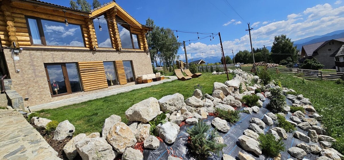 The exterior features a wooden cabin with large windows, surrounded by a landscaped yard. A rocky stream flows through the foreground, complemented by green vegetation. Expansive grass areas are paired with lounge chairs, set against a backdrop of distant hills and a clear blue sky.